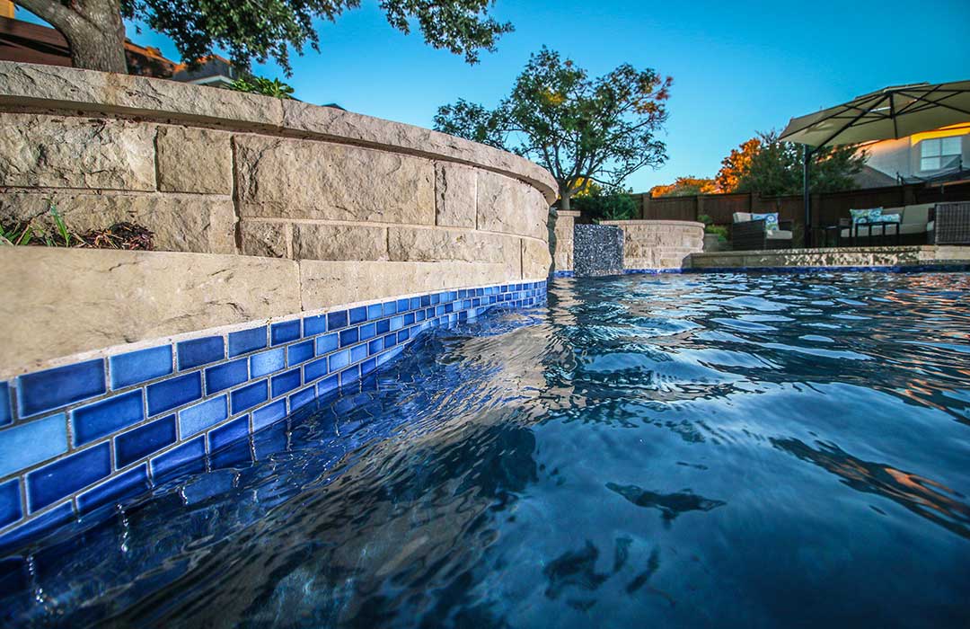 Pool with blue tiled edge and stone wall, surrounded by trees and a building.