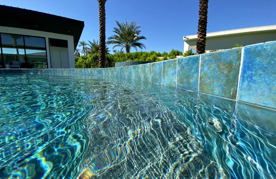 Clear blue pool water with blue waterline tile, palm trees, and a clear sky.