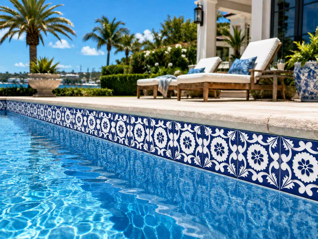 Blue and white patterned ceramic pool tiles lining a swimming pool next to a patio with lounge chairs, palm trees, and a house in the background.