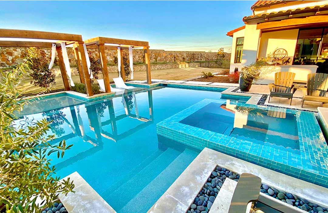 Outdoor pool area with blue tile on spa, lounge chairs, and patio furniture on a sunny day.